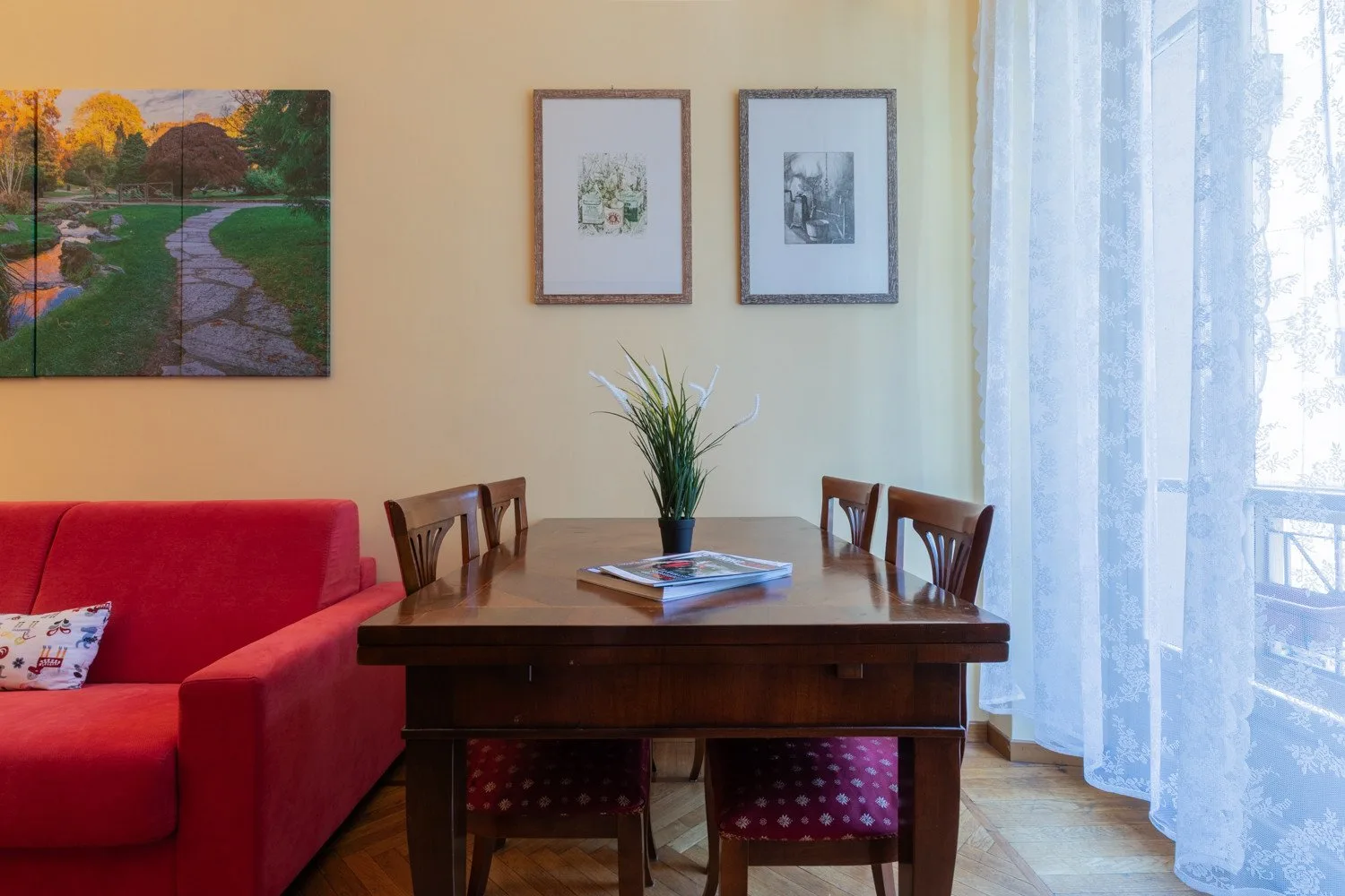 Una mesa de comedor con asientos está junto a un sofá rojo y una ventana luminosa con cortinas.