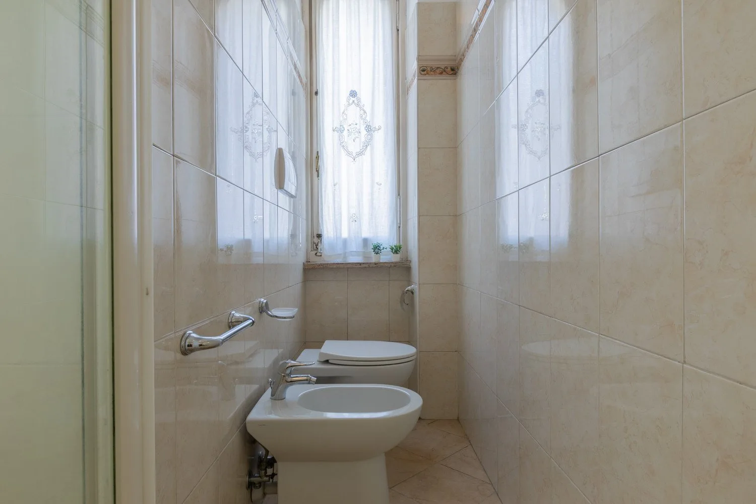Un baño alicatado con inodoro y lavabo bajo una ventana con cortina.