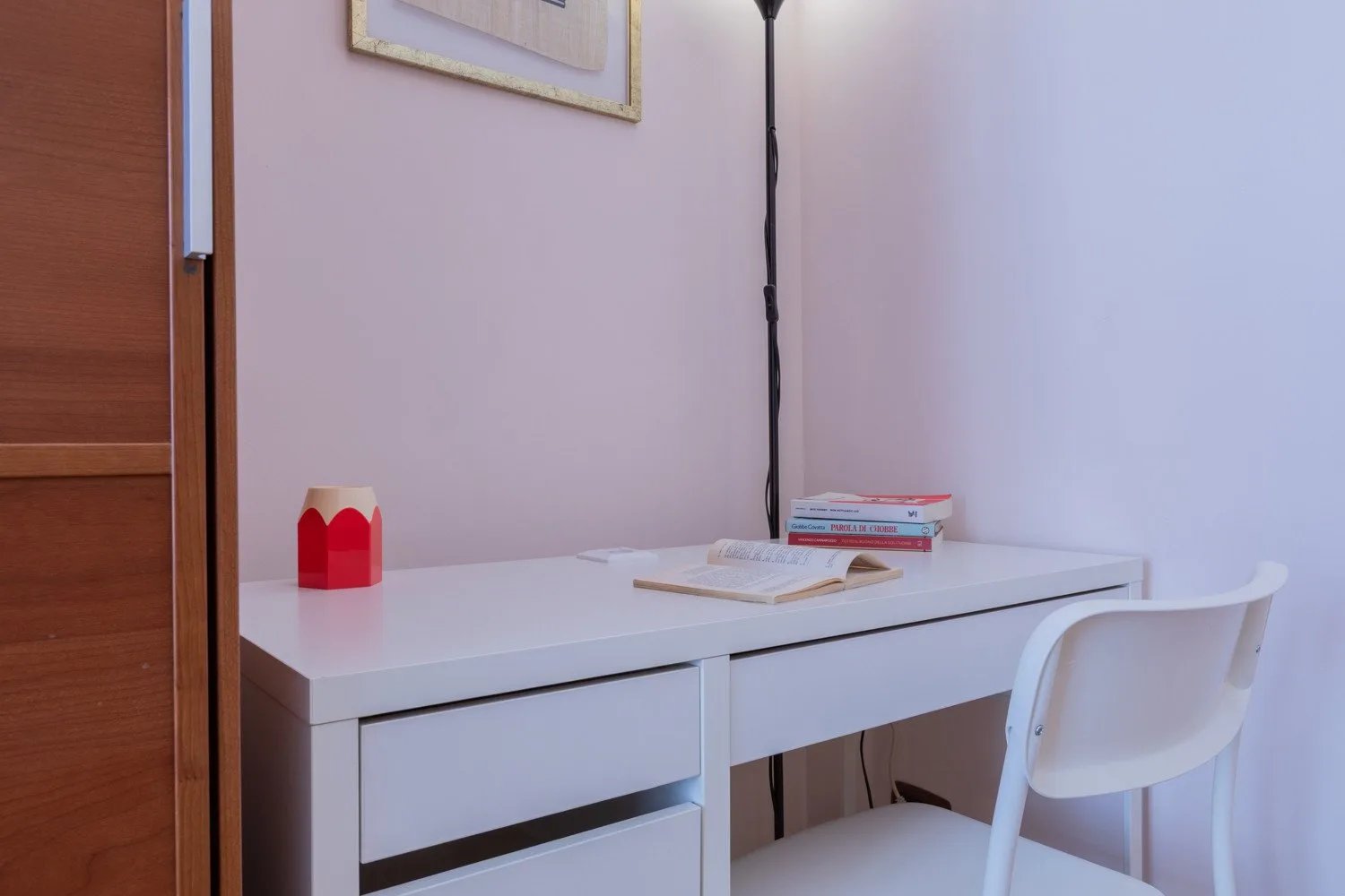 Un escritorio blanco y una silla están junto a una pared clara, con libros sobre el escritorio.