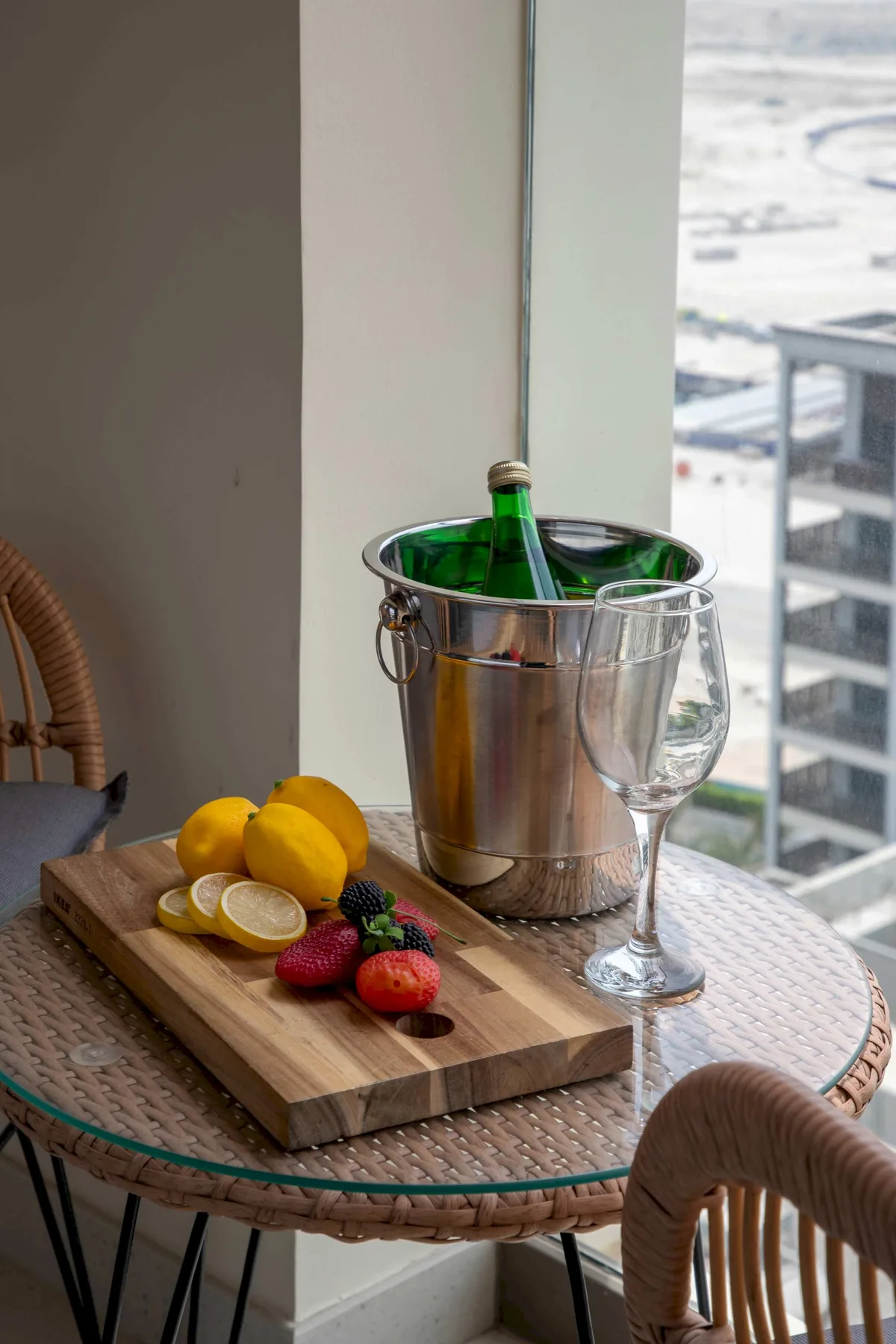 Una mesa junto a una ventana tiene una cubitera, una copa y fruta.