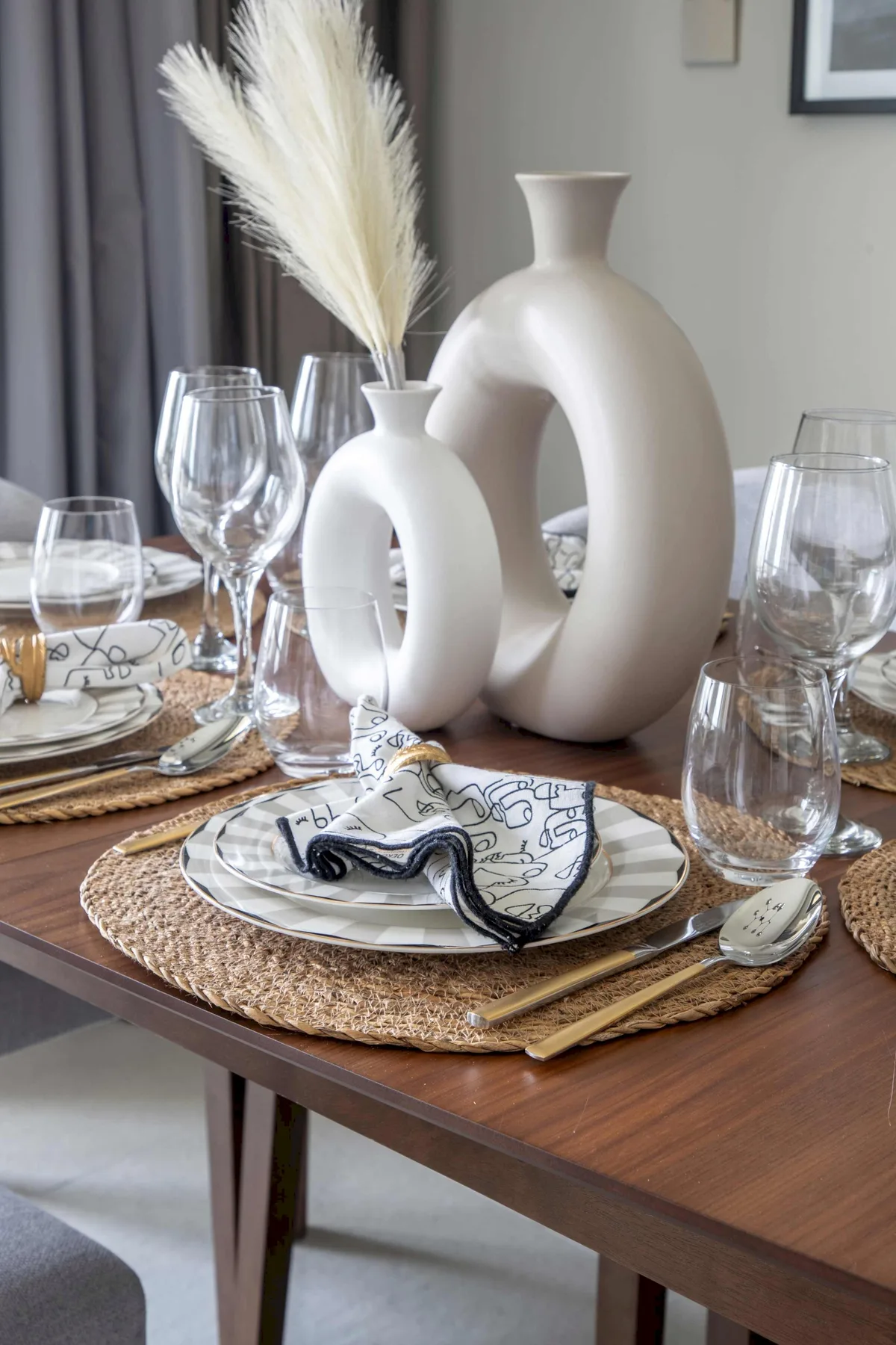 Una mesa de comedor está puesta con platos, copas, manteles individuales tejidos y jarrones decorativos.