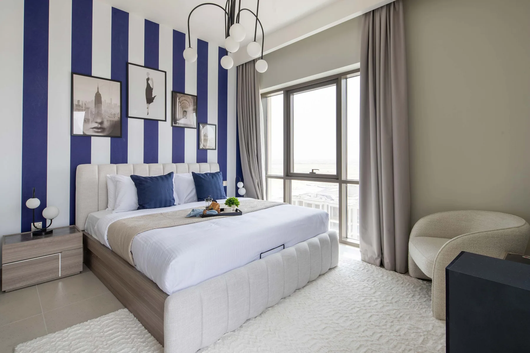 Un dormitorio luminoso con una cama tendida, una pared a rayas y ventanas altas.