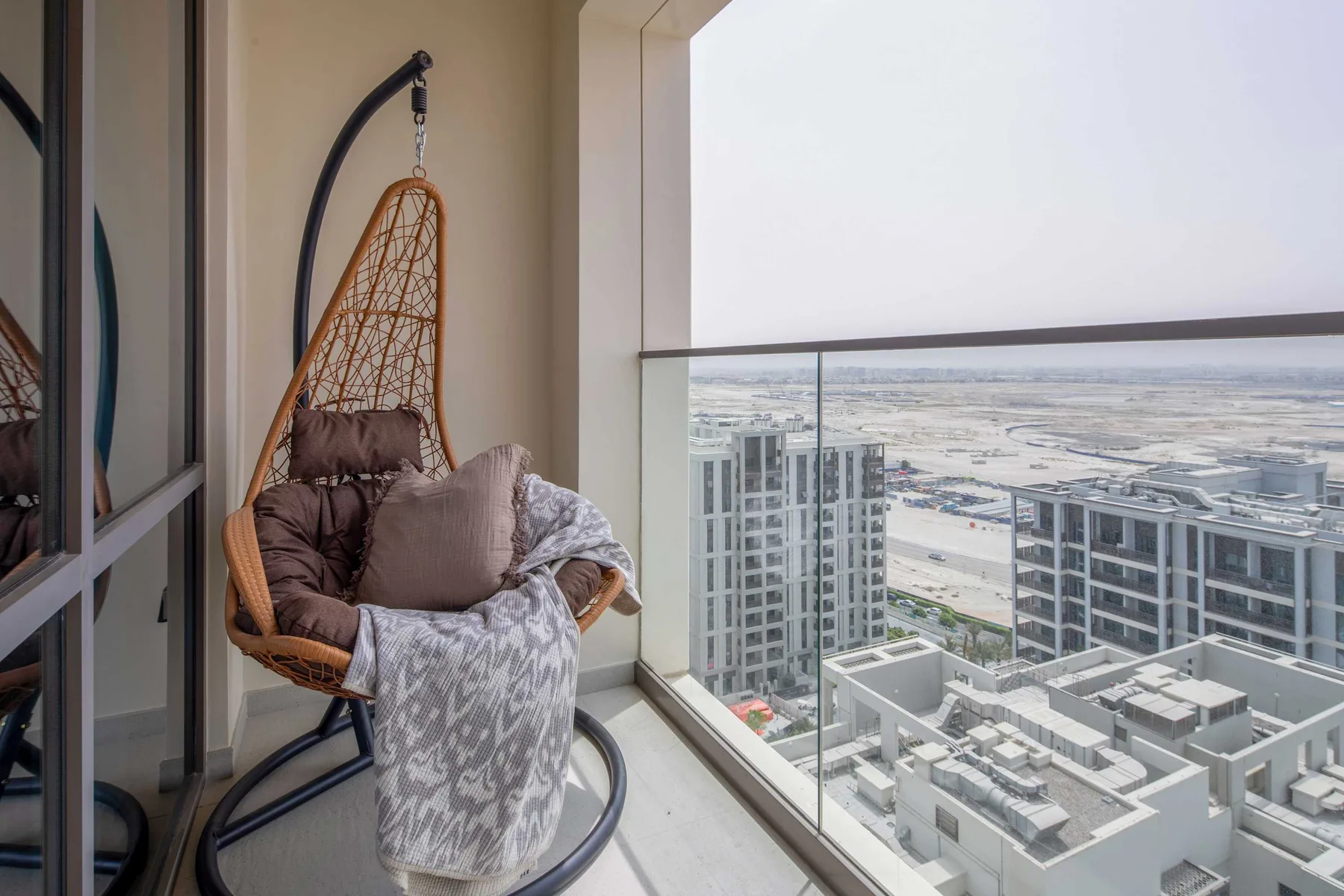 Zona de estar en un balcón con una silla colgante junto a una barandilla de vidrio, con vistas a la ciudad.