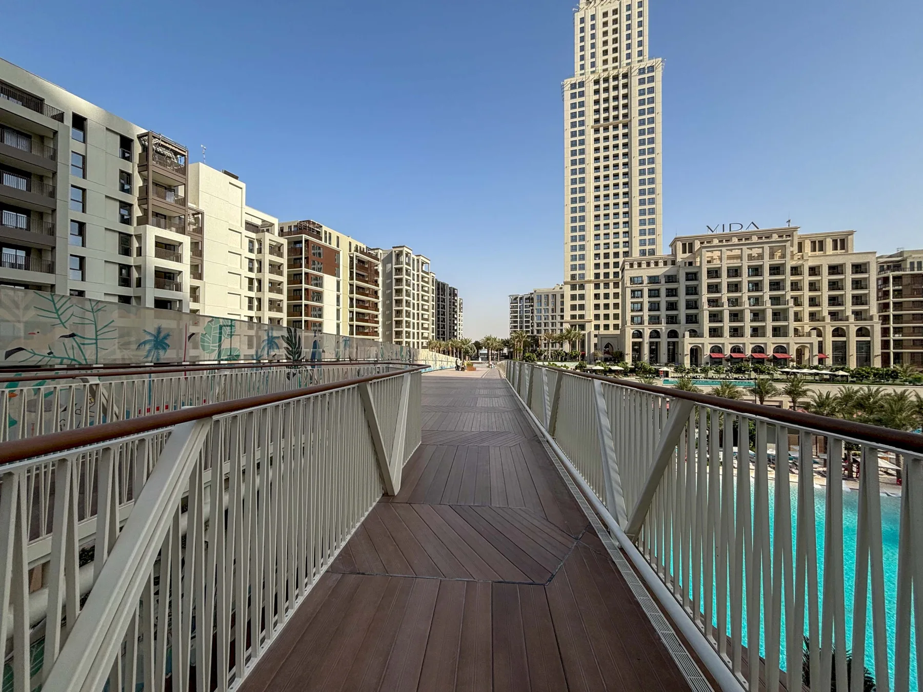 Una pasarela peatonal con barandillas cruza sobre una piscina turquesa entre edificios de apartamentos.