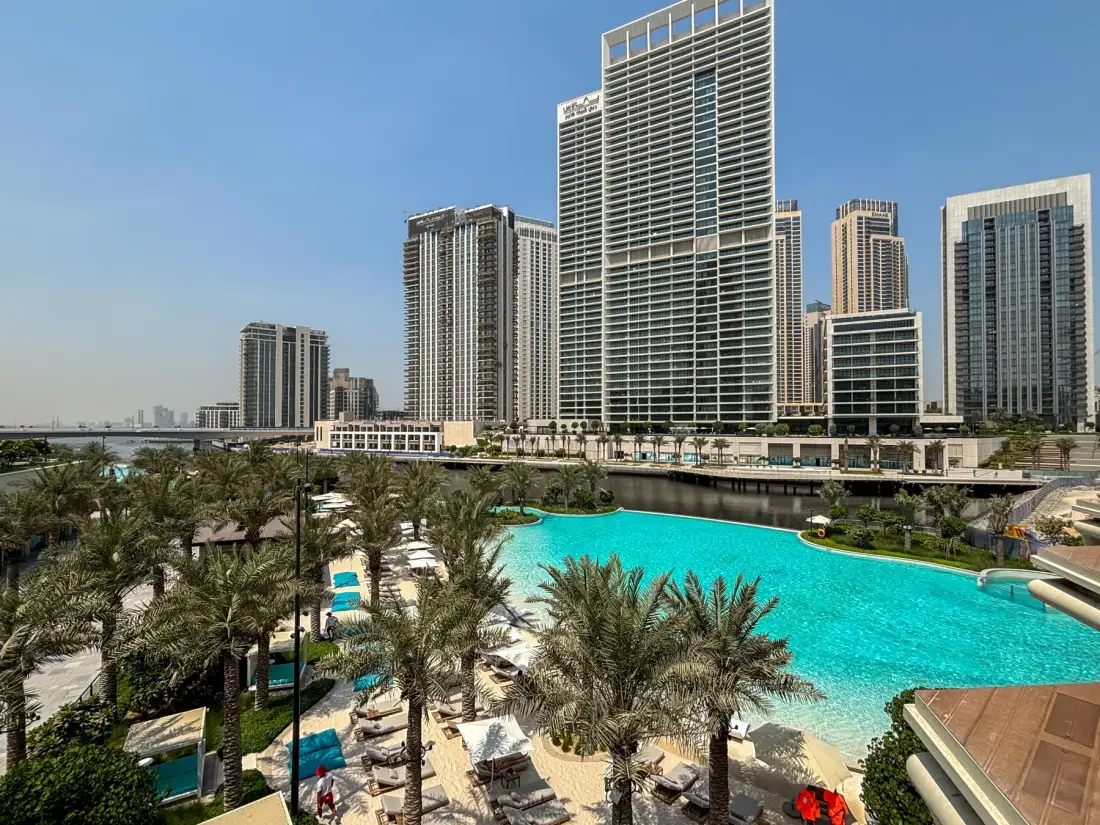 Una piscina en una terraza está rodeada de palmeras, con edificios altos al fondo.