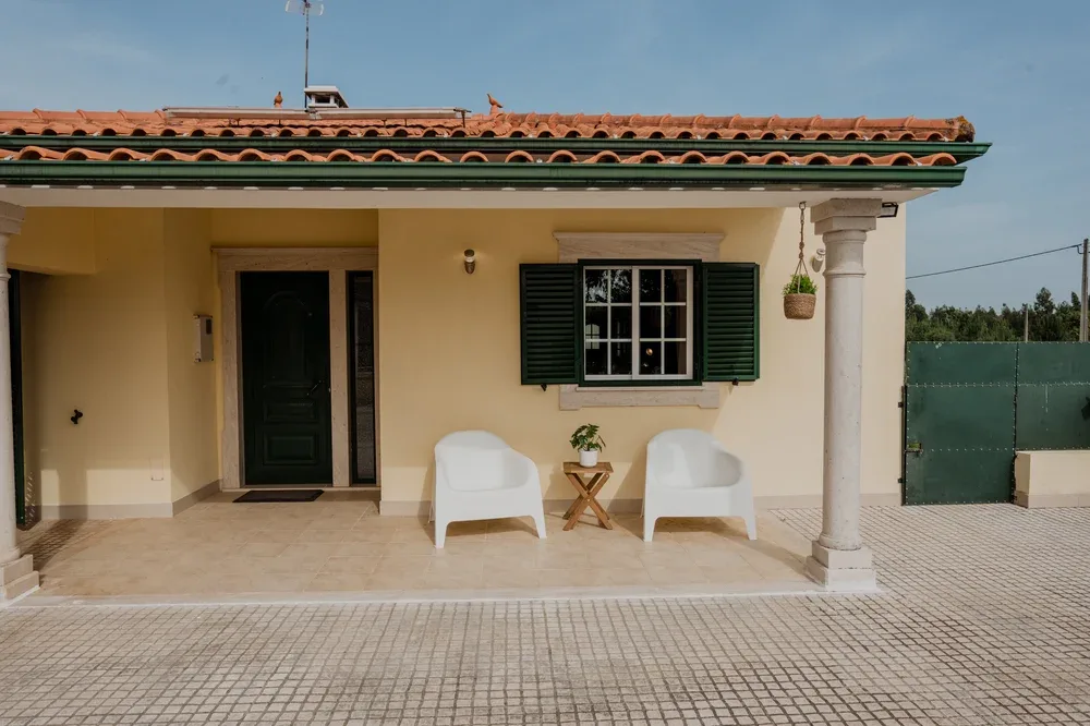 Una entrada de casa con dos asientos blancos junto a una ventana con contraventanas verdes.