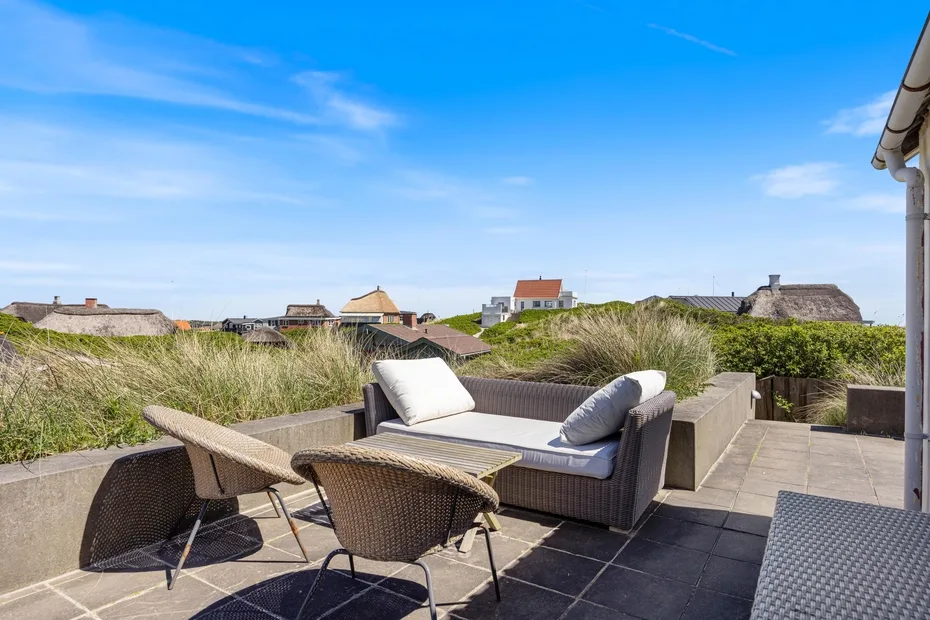 Un patio al aire libre tiene asientos acolchados y vistas a dunas cubiertas de hierba y casas cercanas.