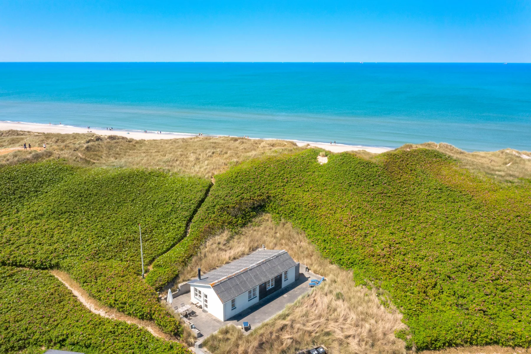 Una casa se encuentra entre dunas costeras, con la playa y el mar al fondo.
