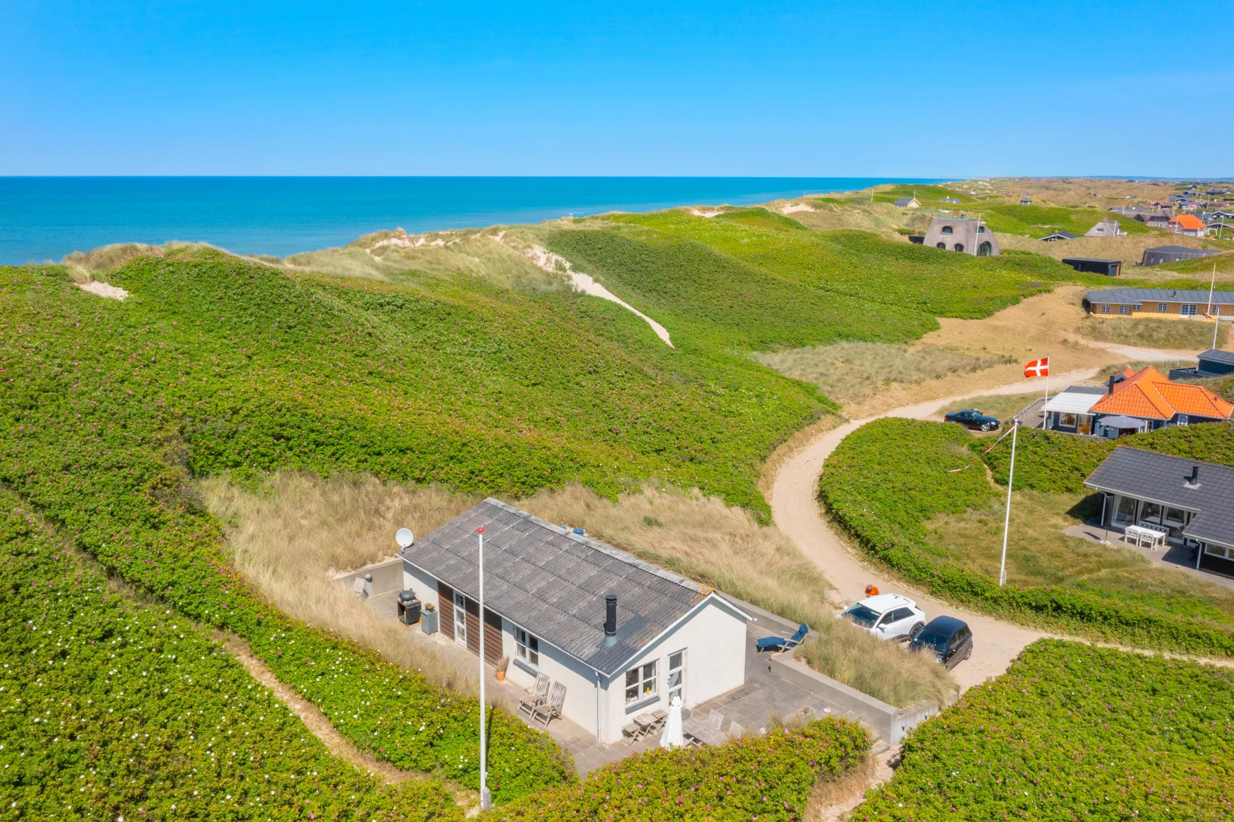 Una casa costera se encuentra entre dunas verdes junto a un camino de arena curvo.