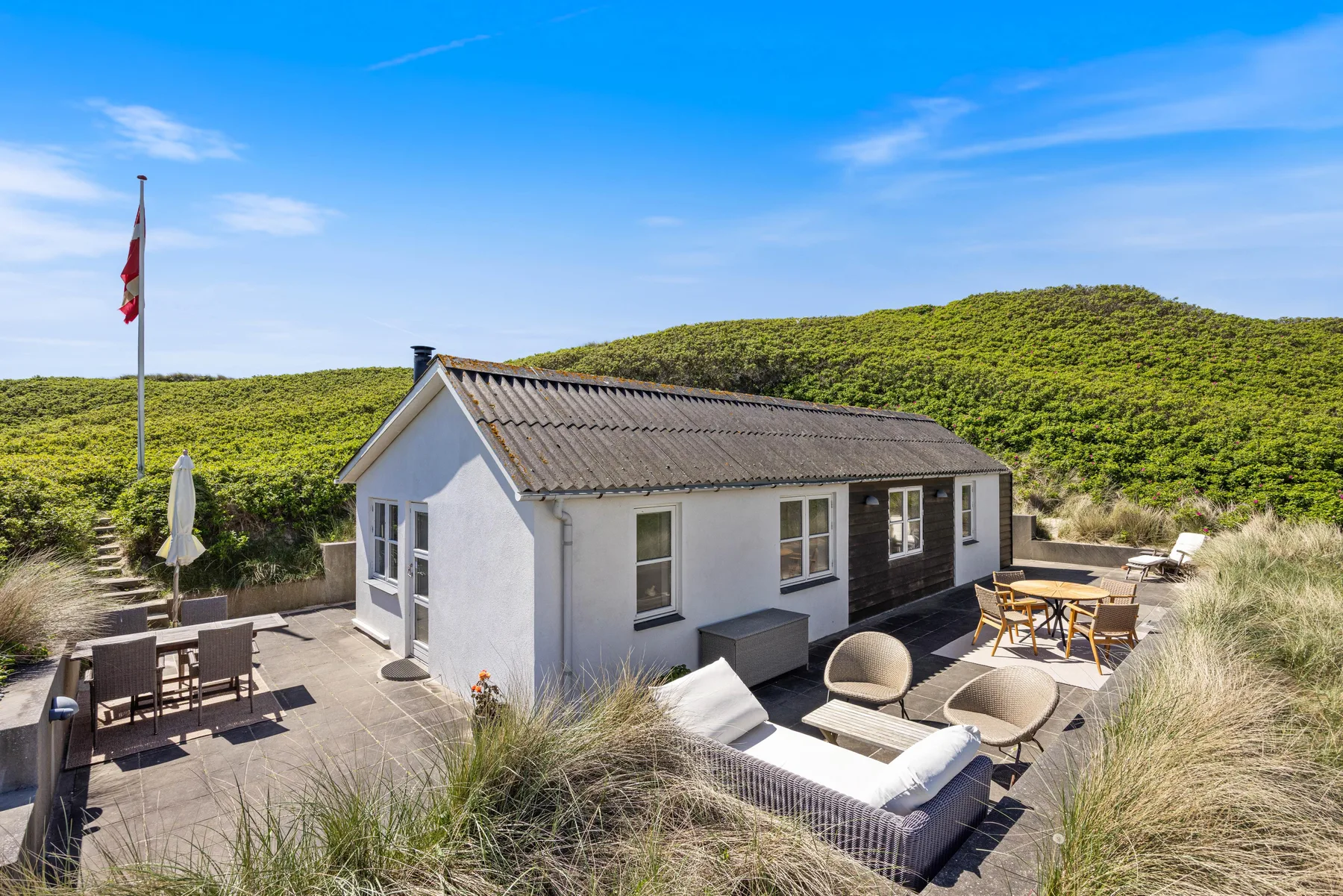Una casa blanca con comedor al aire libre se encuentra junto a dunas cubiertas de hierba bajo un cielo azul.