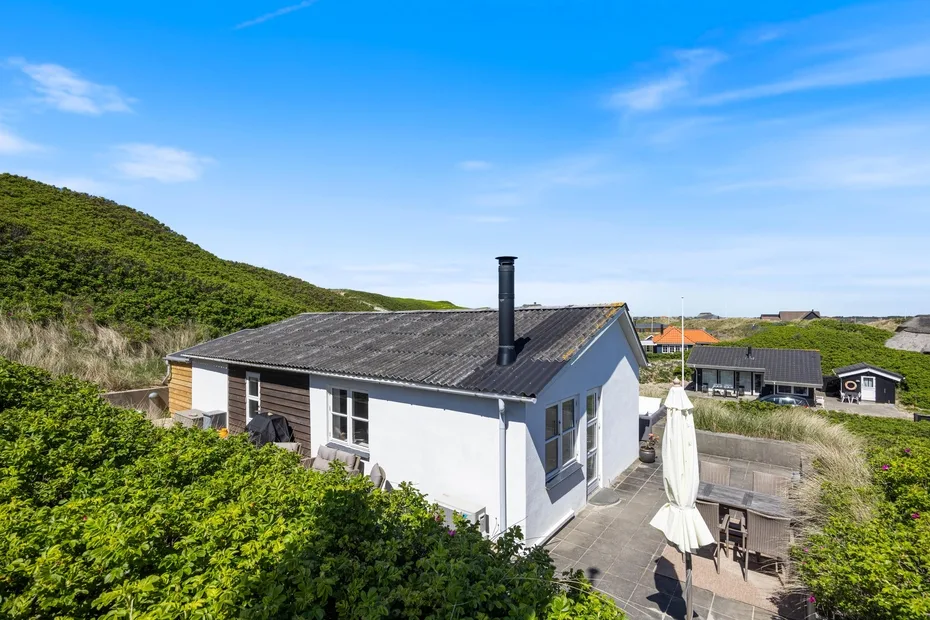 Una casa blanca con techo oscuro junto a un patio y una ladera verde.