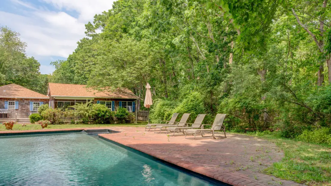 An outdoor swimming pool beside a paved patio with lounge seating and trees.