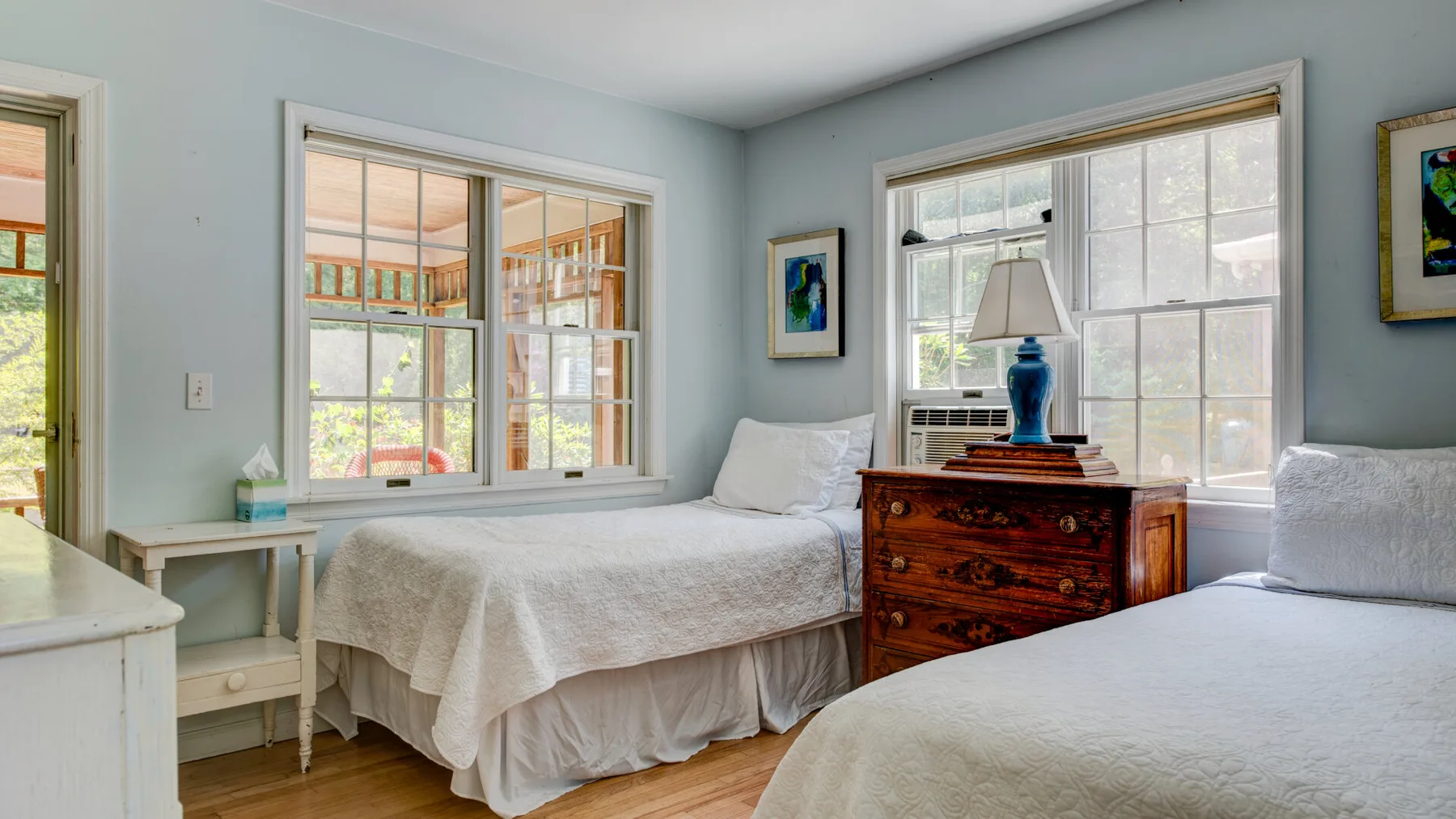 A bright bedroom with two beds, large windows, and a wooden dresser.
