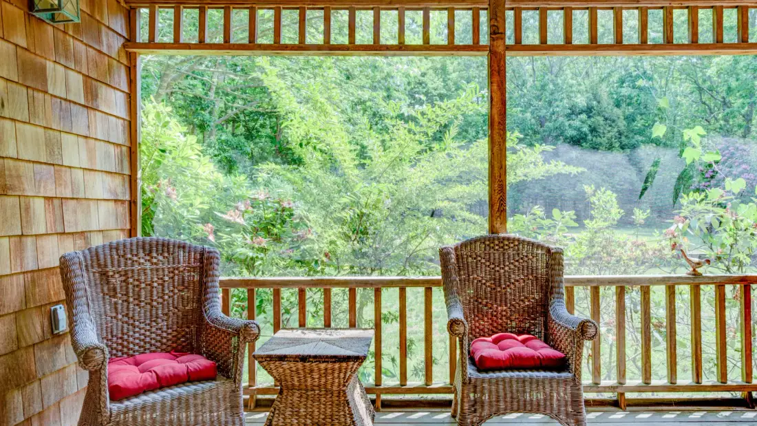A covered seating area with two wicker chairs and a leafy view through large windows.