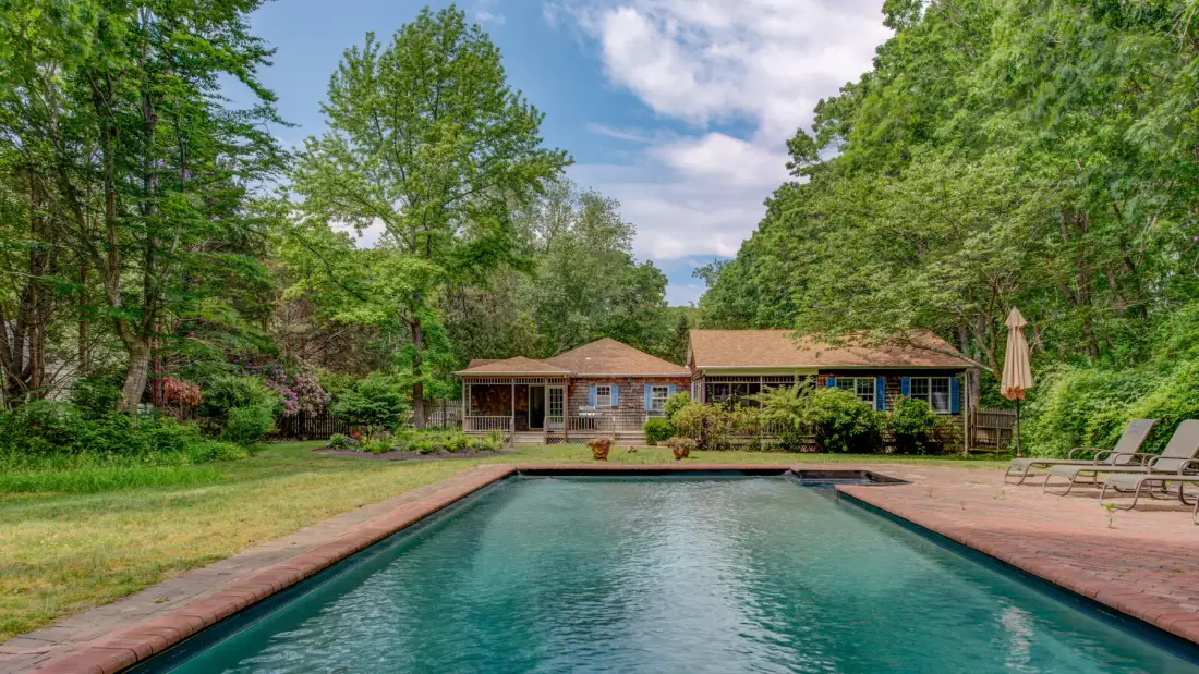 An outdoor swimming pool sits in front of a low house surrounded by trees.