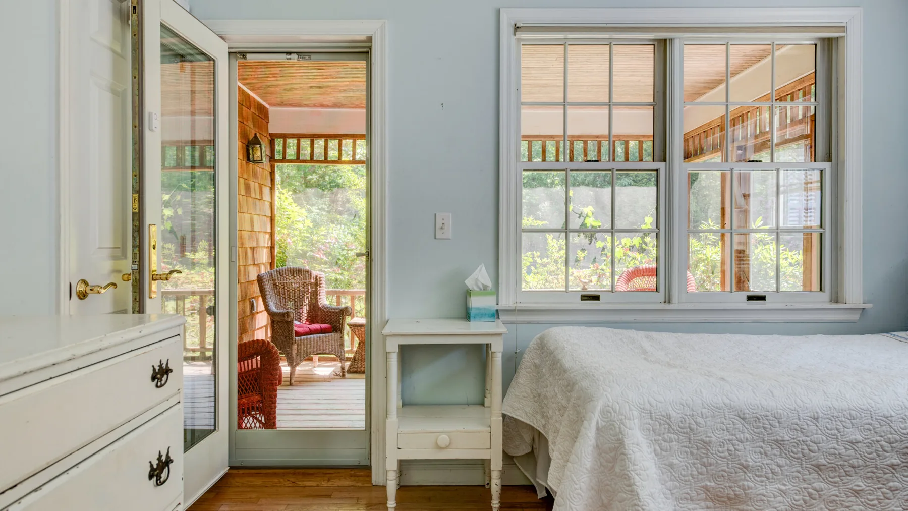A bedroom with a bed beside a window overlooking a sunlit porch.