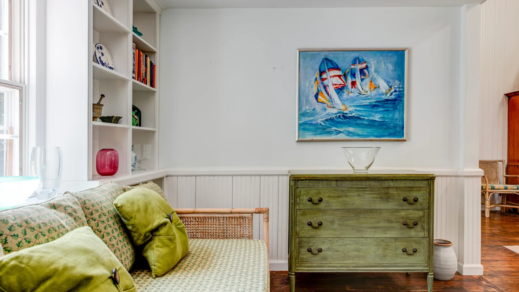A bright sitting room with cushioned seating and a green dresser beneath a sailboat painting.