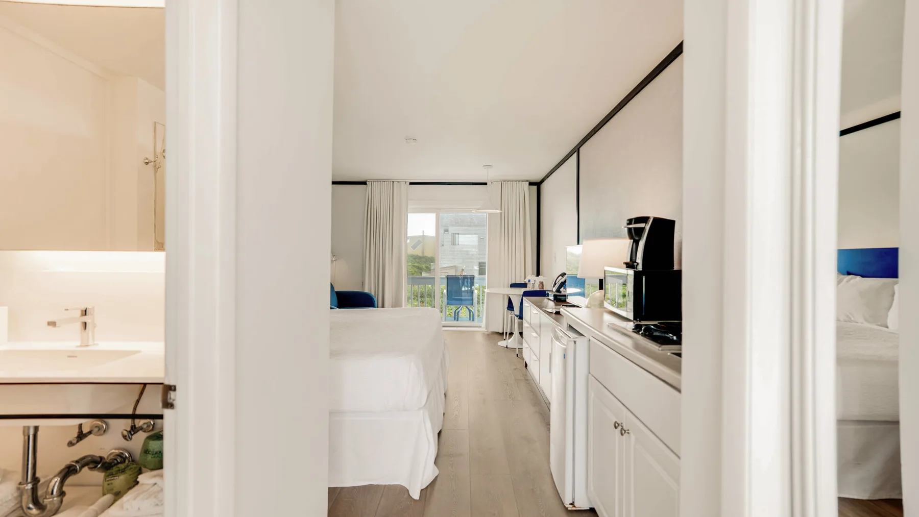 Un dormitorio luminoso da a una pequeña cocina, con una cama y armarios junto al pasillo.