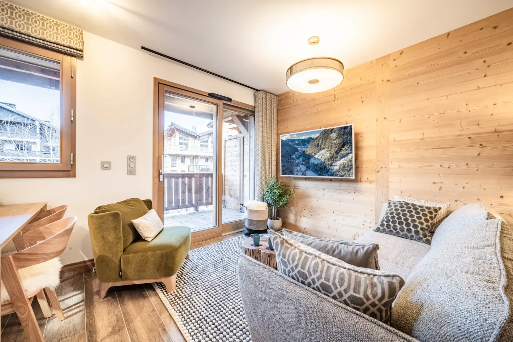 Cozy living room with a green armchair, grey sofa, wall-mounted TV, and wooden walls. There's a dining area to the left and a glass door leading to a balcony with views of buildings outside.