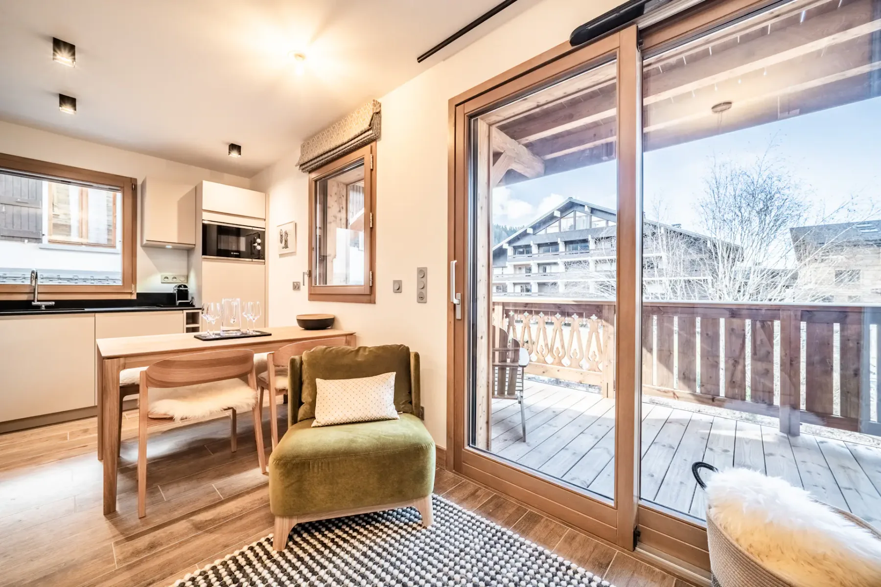 Cozy living room with a green armchair, grey sofa, wall-mounted TV, and wooden walls. There's a dining area to the left and a glass door leading to a balcony with views of buildings outside.