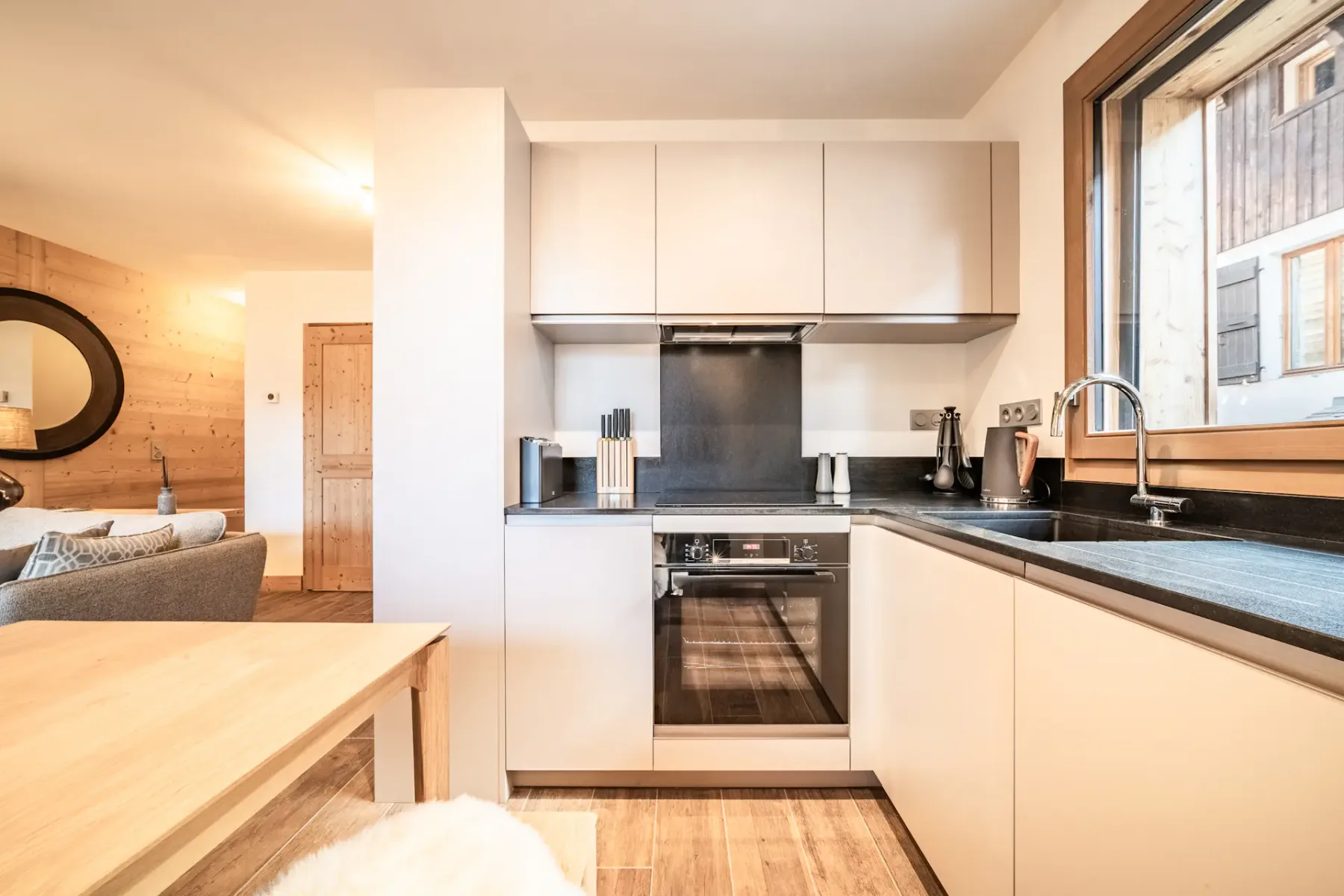 Modern kitchen and dining area with wooden table, chairs with fur cushions, and large windows. Cabinets and appliances are built-in, and a tray with glasses is on the table.