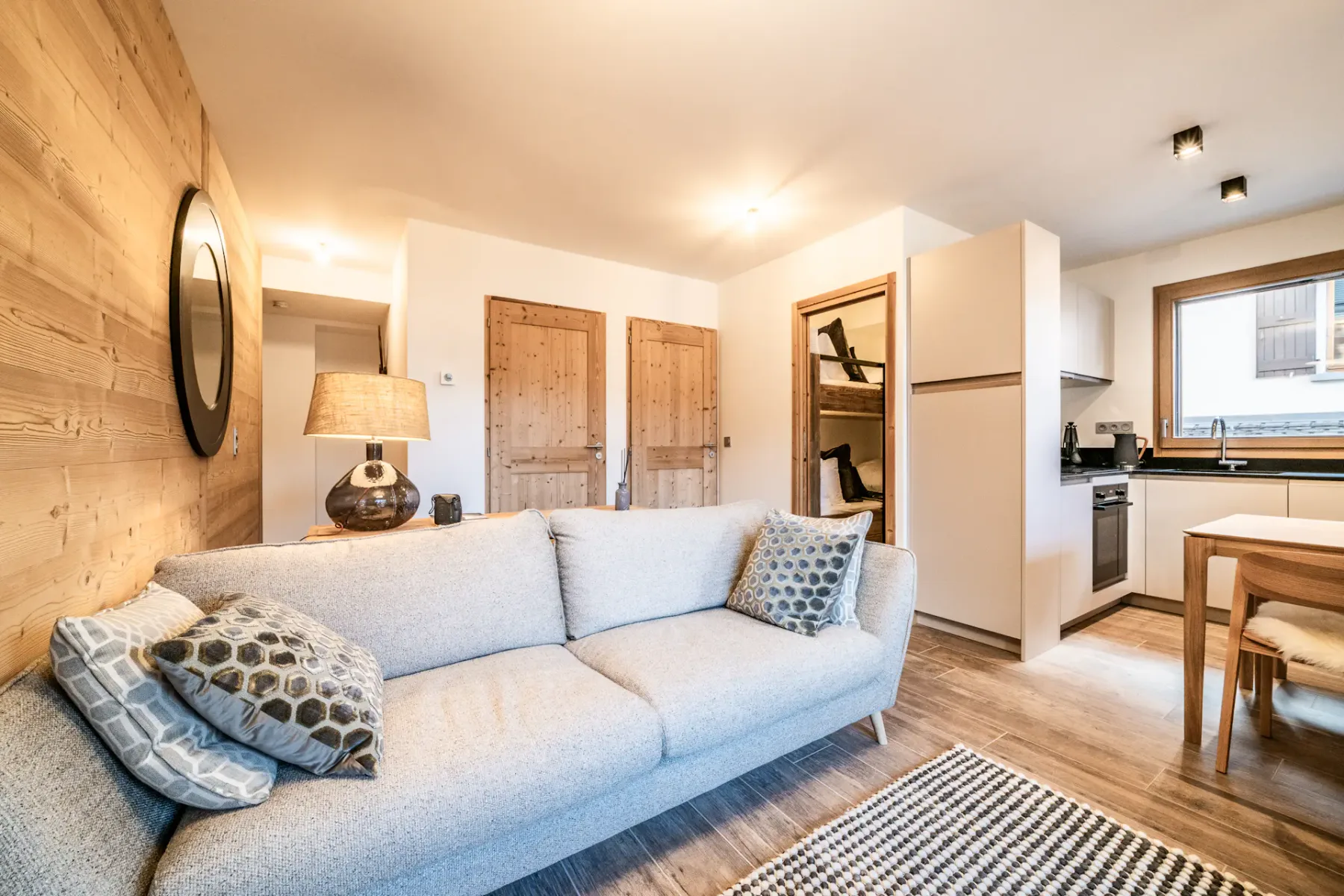 Sala de estar acogedora con un sillón verde, un sofá gris, un televisor montado en la pared y paredes de madera. A la izquierda hay una zona de comedor y una puerta de cristal que da a un balcón con vistas a los edificios del exterior.