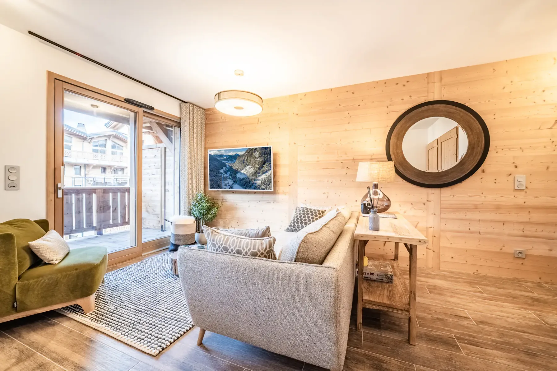 Cozy living room with a green armchair, grey sofa, wall-mounted TV, and wooden walls. There's a dining area to the left and a glass door leading to a balcony with views of buildings outside.