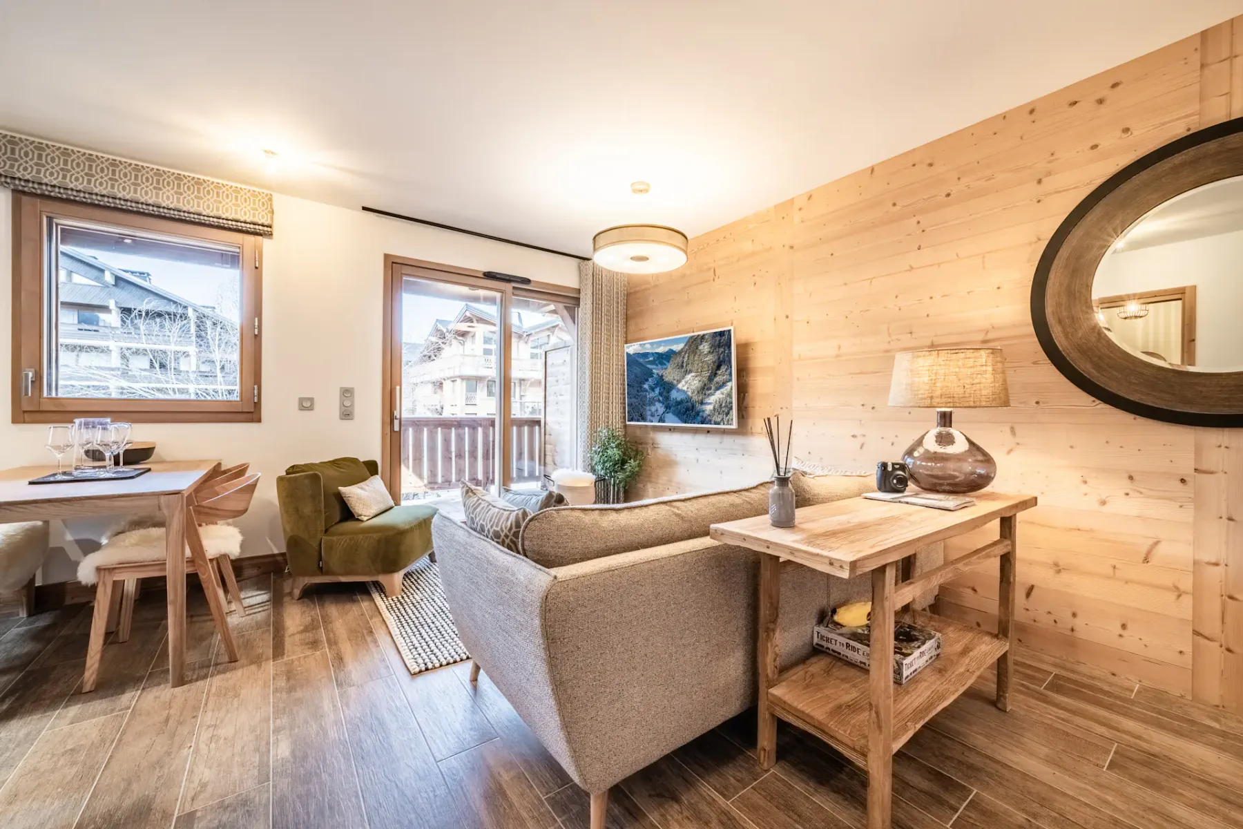Cozy living room with a green armchair, grey sofa, wall-mounted TV, and wooden walls. There's a dining area to the left and a glass door leading to a balcony with views of buildings outside.