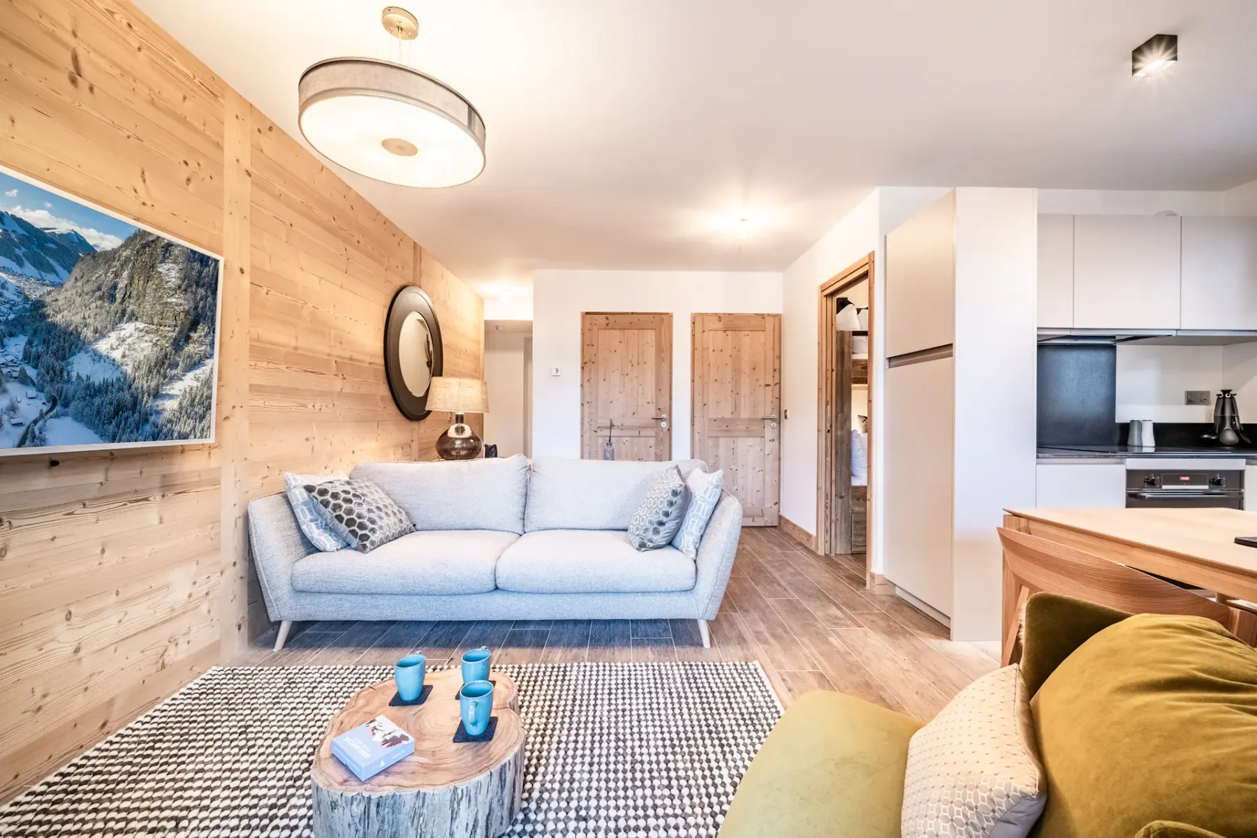 Cozy living room with a green armchair, grey sofa, wall-mounted TV, and wooden walls. There's a dining area to the left and a glass door leading to a balcony with views of buildings outside.