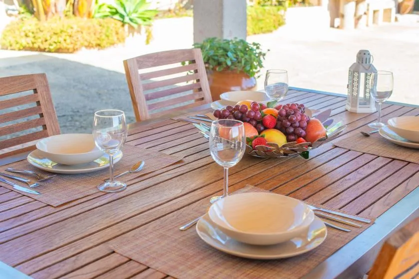 An outdoor dining table is set with dishes, glasses, and a bowl of fruit.