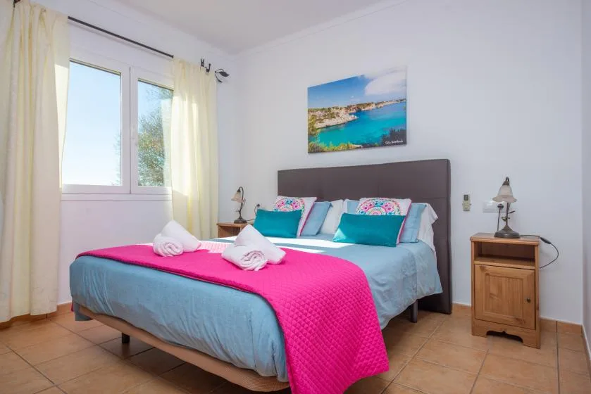 A bedroom with a bed dressed in bright pink and blue bedding beside a window.