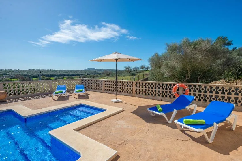 A sunny pool terrace with blue lounge seating and an umbrella overlooking green countryside.