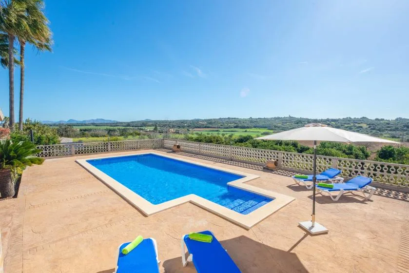 An outdoor swimming pool sits on a sunlit terrace overlooking green fields.