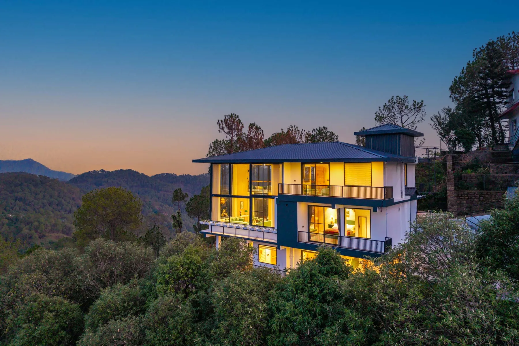 A modern hillside house glows with warm light at dusk, surrounded by trees.