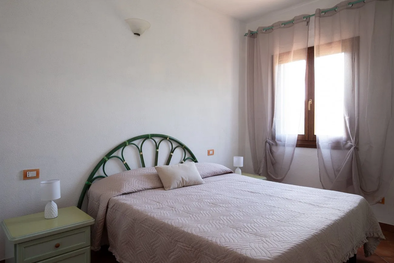 A bedroom with a bed beneath a window with sheer curtains.