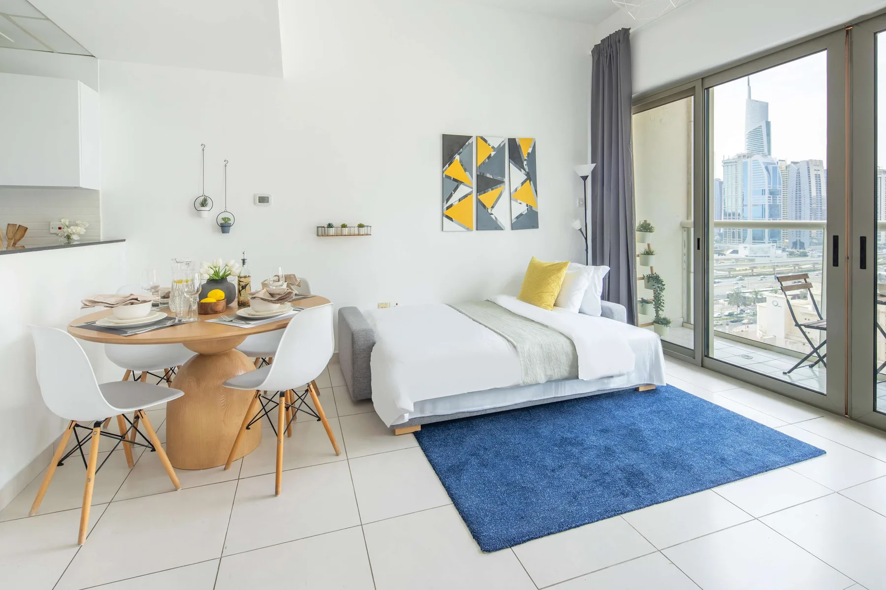 A bright studio room with a dining table, bed, and large windows overlooking the city.