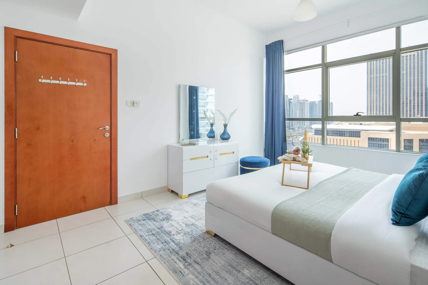 A bright bedroom with a bed beside a large window overlooking nearby buildings.