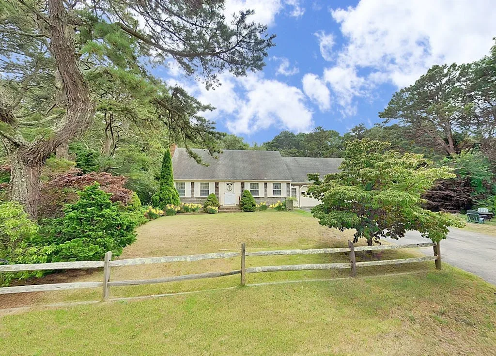 A low house sits behind a wooden fence, surrounded by trees and lawn.