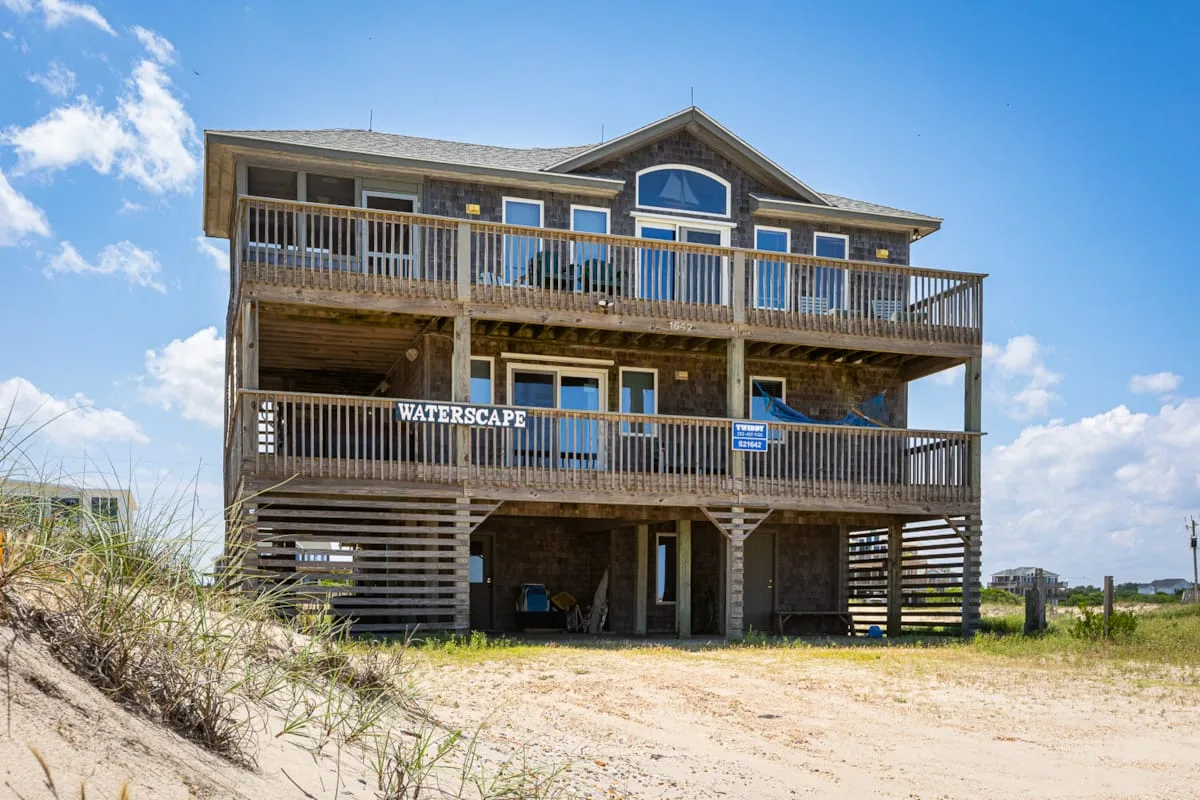 Una gran casa junto a la playa tiene dos balcones de madera sobre una planta baja abierta.