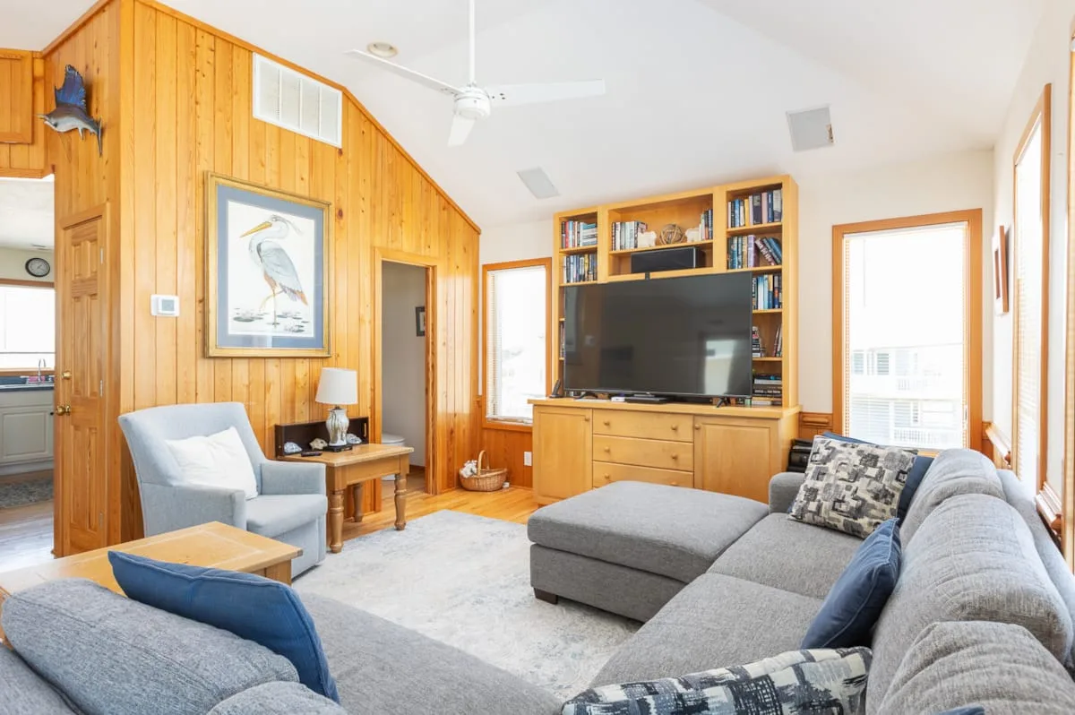 Una sala luminosa con televisor, estanterías de madera y asientos grises.