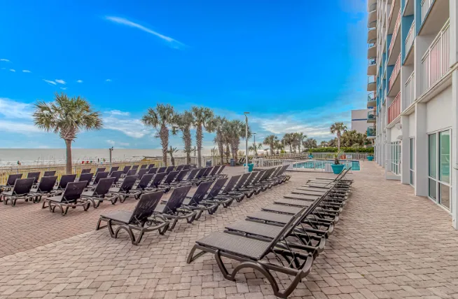Outdoor pool deck with rows of lounge chairs beside a beachfront building.