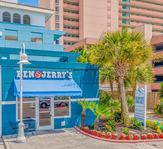 A blue ice cream shop stands beside palm trees and a landscaped flower bed.