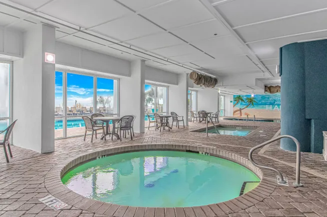 An indoor pool area with a round pool, tiled deck, and tables by the windows.