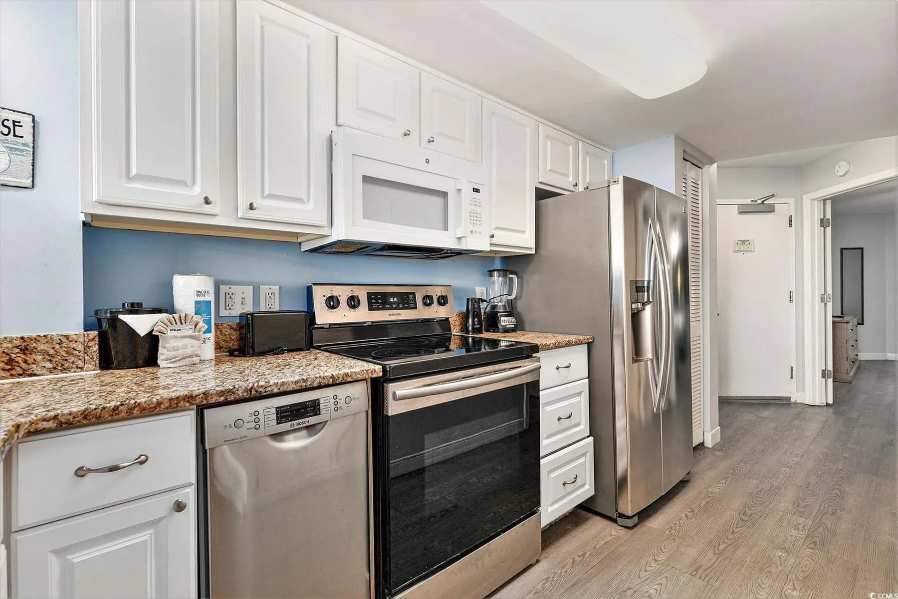 A bright kitchen with white cabinets, stainless steel appliances, and a long countertop.