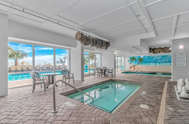 An indoor pool area with a smaller pool in the foreground and windows overlooking an outdoor pool.