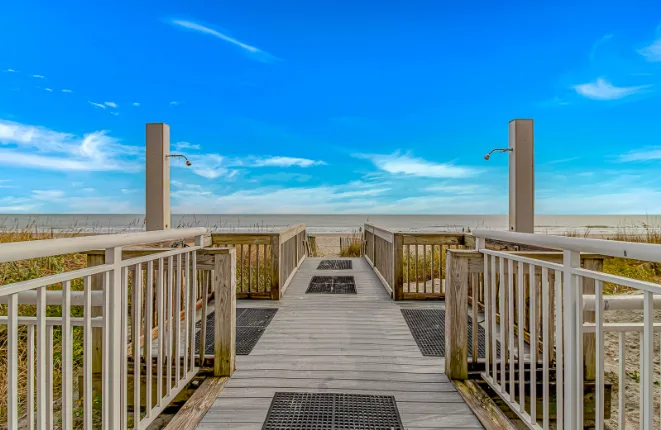 A raised wooden walkway with railings leads toward a sandy beach and open water.