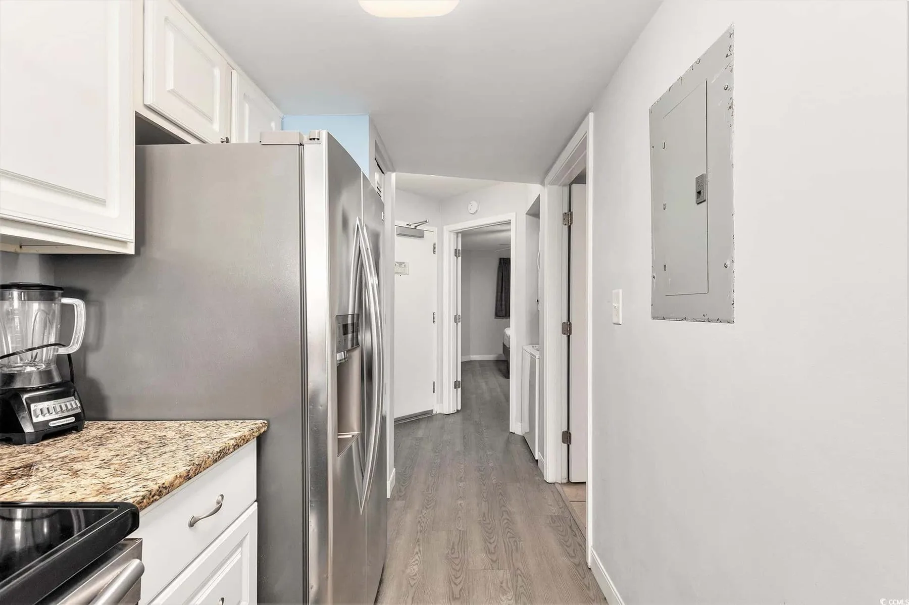 A narrow kitchen opens onto a hallway, with stainless steel appliances and white cabinets.
