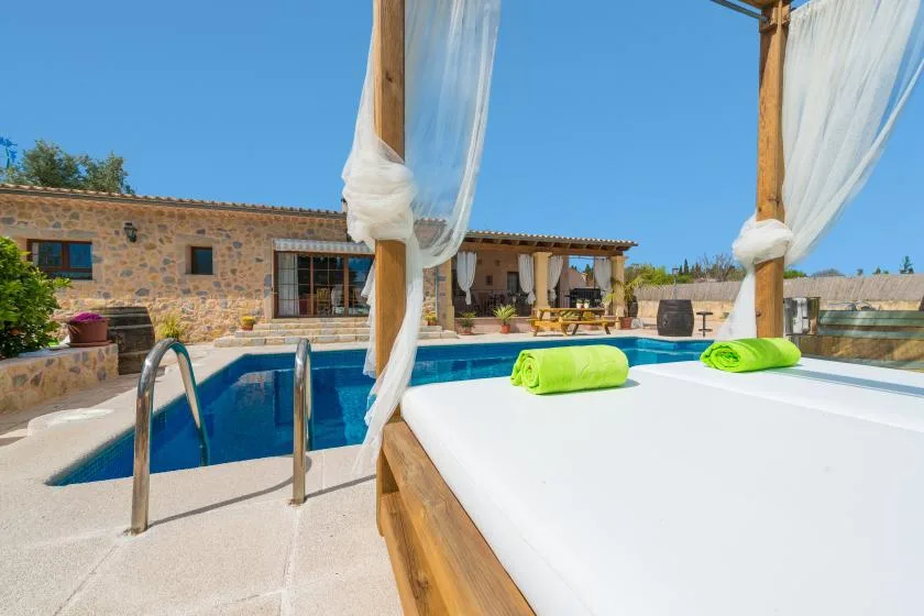 Una zona junto a la piscina con una cama blanca y una casa de piedra bajo un cielo despejado.