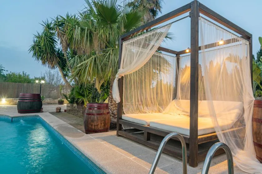 Una cama junto a la piscina está bajo una estructura de madera oscura con cortinas translúcidas.