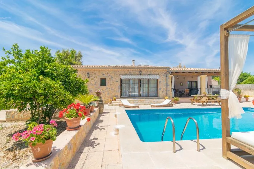 Una piscina exterior soleada junto a una casa de piedra, con plantas floridas junto al patio.